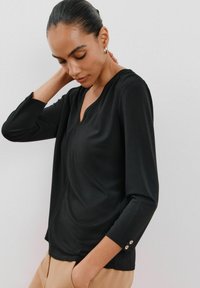 Schwarzes Langarmoberteil mit V-Ausschnitt, weicher Textur und dekorativen Knöpfen an den Bündchen. Lässige Passform, glattes Material und minimalistisches Design.