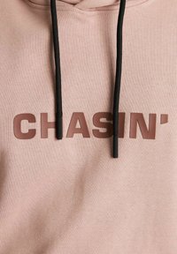 Felpa con cappuccio di un rosa polveroso realizzata in tessuto morbido, con una grafica "CHASIN’" di colore marrone scuro nella parte anteriore e dettagli con cordini neri.
