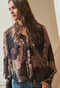 Kerge must blouse roosa ja rohelise lillemustriga, millel on sidumisnööriga kaelus ja volangis varrukad, kombineerituna siniste teksadega.