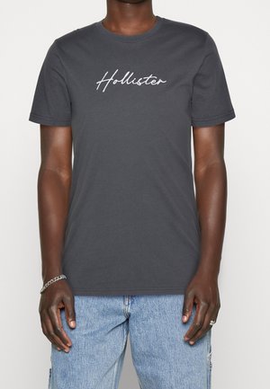 Ciemnoszary T-shirt z krótkim rękawem z napisem "Hollister", noszony przez osobę o średnio ciemnej karnacji i jasnoniebieskich dżinsach.