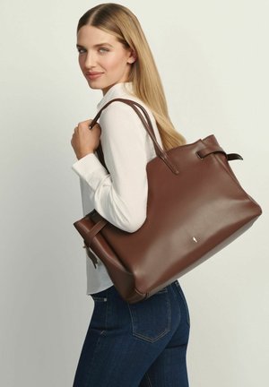 Kobieta o długich blond włosach, ubrana w białą bluzkę i dżinsy, niesie na ramieniu dużą brązową skórzaną torbę typu tote.
