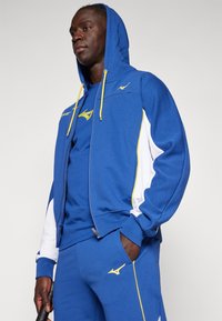 Hanorac albastru cu fermoar, cu accente galbene și panouri albe, asortat cu pantaloni de sport albastre. Dispune de glugă și detalii ale logo-ului vizibile.