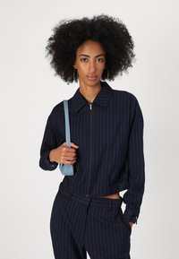 Marineblauwe gestreepte blazer met een kraag en een rits aan de voorkant, met een cropped pasvorm, textuurstof en relaxte mouwen, gecombineerd met een lichtblauwe tas.