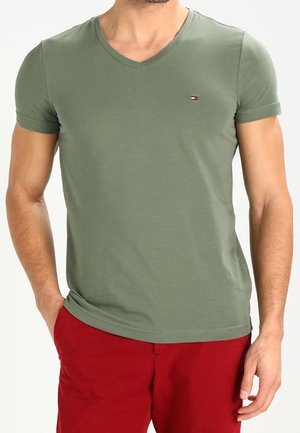 Man draagt olijfgroene V-hals T-shirt met klein logo op de borst en rode korte broek, handen ontspannen langs het lichaam.