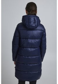 Marineblauwe pufferjas met capuchon, met een geborduurde textuur, lange lengte en horizontale stiksels, voorzien van een gladde, glanzende afwerking.