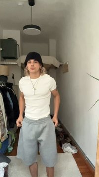 Tricou alb cu mânecă scurtă, pantaloni scurți de trening gri, în picioare în interior. Beanie negru, pandantiv argintiu și accente din lemn deschis în fundal.