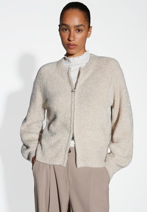 Vrouw in een lichtbeige fuzzy zip-up cardigan over een witte geribbelde coltrui, ingestopt in hoge taille taupe wijde broek.