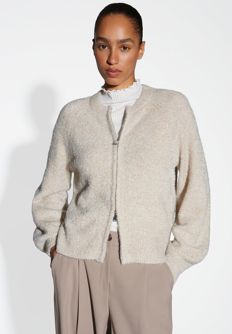 Vrouw in een lichtbeige fuzzy zip-up cardigan over een witte geribbelde coltrui, ingestopt in hoge taille taupe wijde broek.