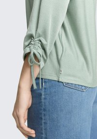 Zeleni prugasti dugih rukava s naboranim rukavima s vezicama i ležernim krojem, upareni s plavim trapericama. Vidljiva teksturirana tkanina.