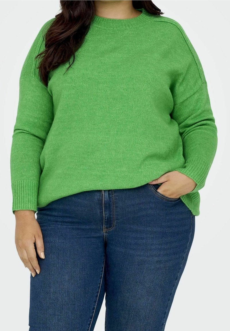 Femme portant un pull vert vif à manches longues et un jean bleu foncé, une main dans la poche, sur un fond clair uni.