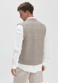 Gestrickte Weste in Hellgrau mit Fischgrätmuster, getragen über einem weißen Langarmshirt, mit geripptem Saum und ärmellosem Design.