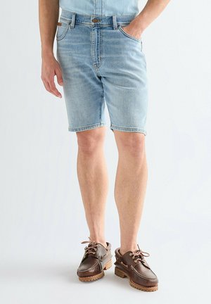 Hombre con pantalones cortos de mezclilla azul claro y zapatos náuticos de cuero marrón, de pie con una mano en el bolsillo.