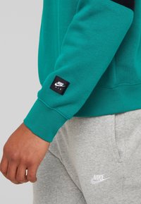 Grön sweatshirt med svarta detaljer och en strukturerad tyg. Har en svart Nike Air-tag på ärmen och grå sweatpants med en vit logotyp.