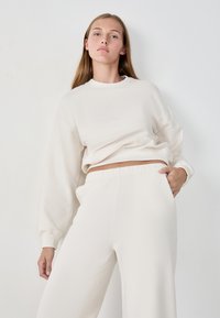 Sudadera color crema con escote redondo y mangas largas, combinada con pantalones anchos a juego. Tejido suave con un ligero brillo.