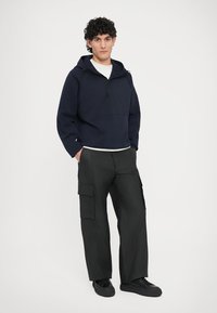 Μπλε ναυτικού χρώματος φούτερ με φερμουάρ και κουκούλα, με υφή, συνοδευόμενο από μαύρο, φαρδύ παντελόνι cargo με πλαϊνές τσέπες και ματ υφή.