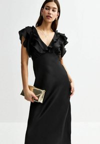 Abito maxi nero con scollatura profonda a V, maniche corte a palloncino e una texture liscia. La modella tiene in mano una pochette metallica dorata.