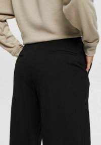 Svarta byxor med en avslappnad passform, med en mjuk textur och två bakfickor. Matcher med en beige sweatshirt som delvis syns.