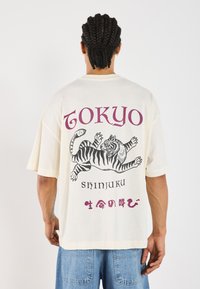 Túlsúlyos krémszínű póló fekete-fehér tigris grafikával. Lila színű szöveg olvasható rajta: "TOKYO" és "SHINJUKU", alatta japán karakterekkel.