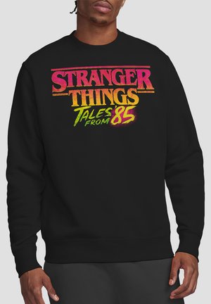 Mann trägt schwarzen Pullover mit farbigem Schriftzug "Stranger Things Tales from 85" vorne und steht vor einfachem Hintergrund.