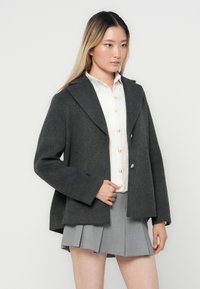 Blazer en mélange de laine gris avec un col cranté et des boutons doubles, porté sur une chemise blanche à boutons et une jupe plissée grise.