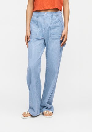 Γυναίκα που φορά φαρδιές παντελόνες με ανοιχτό μπλε και λευκές ρίγες, με μεγάλες μπροστινές τσέπες και λευκά σαγιονάρα, σε συνδυασμό με ένα κοντό πορτοκαλί τοπ.