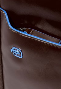 Dettaglio di una borsa in pelle marrone che mostra una vivace rifinitura blu e un piccolo logo blu. Tasca con zip visibile all'interno della borsa. Texture liscia.