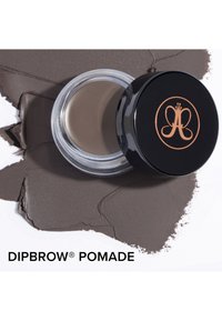 Dipbrow Pomade w przezroczystym słoiczku z czarną pokrywką z złotym logo, otoczony taupe'owym kremem do brwi na białym tle.