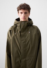 Olivengrüne, leichte, wasserdichte Jacke mit hohem Kragen, verstellbarem Zugband und minimalistischer Gestaltung. Sanfte Textur, keine sichtbaren Muster.