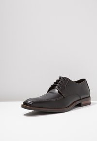 Chaussure habillée en cuir noir avec bout arrondi, fermeture à lacets et surpiqûres contrastantes marron. Dispose d'un petit talon empilé.