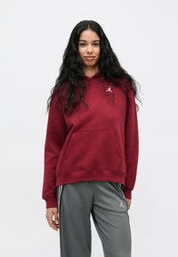 Ung kvinne med langt, krøllete svart hår iført en vinrød hettegenser og grå joggebukser med hvite striper, stående mot en hvit bakgrunn.