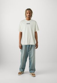 Light green oversized t-shirt featuring black "SHINJUKU" text, paired with light blue baggy jeans and brown sneakers with white accents.