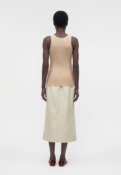 Γυναίκα που στέκεται με την πλάτη, φοράει μπεζ ριγέ χωρίς μανίκια τοπ, κρεμ midi φούστα και σκούρα κόκκινα επίπεδα παπούτσια, σε απλό φόντο.