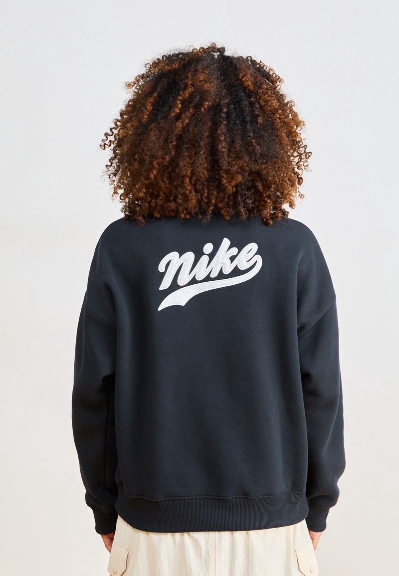 Osoba z kręconymi włosami, ubrana w czarną bluzę z białym logo "nike" na plecach i beżowe spodnie, stoi na jednolitym tle.