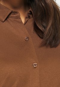 Hnědá košile na knoflíky vyrobená z texturované tkaniny; má límec, jedno knoflíkové zapínání a kontrastní knoflíky.