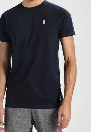 Mand iført en ensfarvet sort kortærmet t-shirt med et lille lyseblåt og hvidt emblem på venstre bryst.