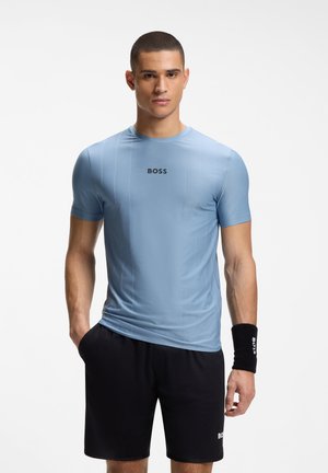Mann trägt hellblaues BOSS-T-Shirt, schwarze Shorts und schwarzes Armband, steht mit einer Hand in der Tasche vor weißem Hintergrund.