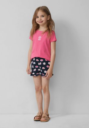 T-shirt di cotone rosa con grafica di conchiglia, abbinato a pantaloni corti blu navy con motivi di conchiglie e volant. Sandali piatti marroni completano il look.
