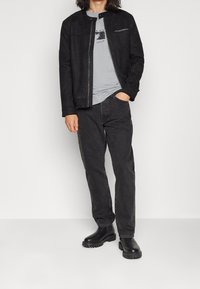 Hombre vestido con una chaqueta de ante negra, una camiseta gráfica gris clara, jeans negros y botas de cuero negras, de pie con una mano en el bolsillo.