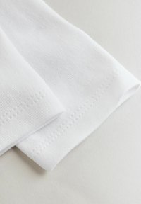 Detail eines weißen Baumwollkleidungsstücks, das die Textur, das gerippte Gewebe und saubere Nähte entlang der Ränder zeigt. Weiches, glattes Aussehen.