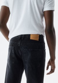 Jeans de mezclilla negra con una etiqueta de cuero en la cinturilla. Presenta dos bolsillos traseros y un corte recto, con detalles de costura visibles.