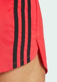 Shorts de sport rouges avec des rayures noires sur les côtés, un détail d'ourlet fendu et une texture lisse. Le design met l'accent sur la mobilité et le confort.