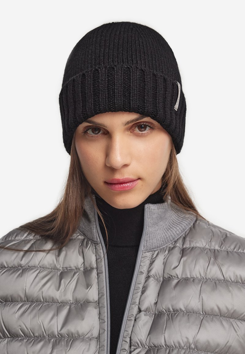 Femme portant un bonnet en tricot noir et une veste matelassée grise avec un col roulé noir en dessous, expression faciale neutre, fond blanc.