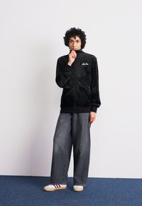 Sorte fløjl zip-up jakke med høj krave, der har et logo på brystet, parret med løse grå denim bukser og hvide sneakers med røde striber.