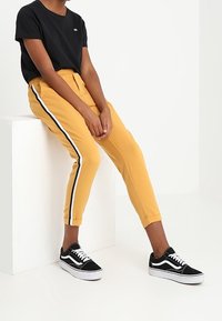 Svart kortärmad T-shirt med logotyp, gula tapered byxor med vita sidostråkar och svarta sneakers med vita accenter.