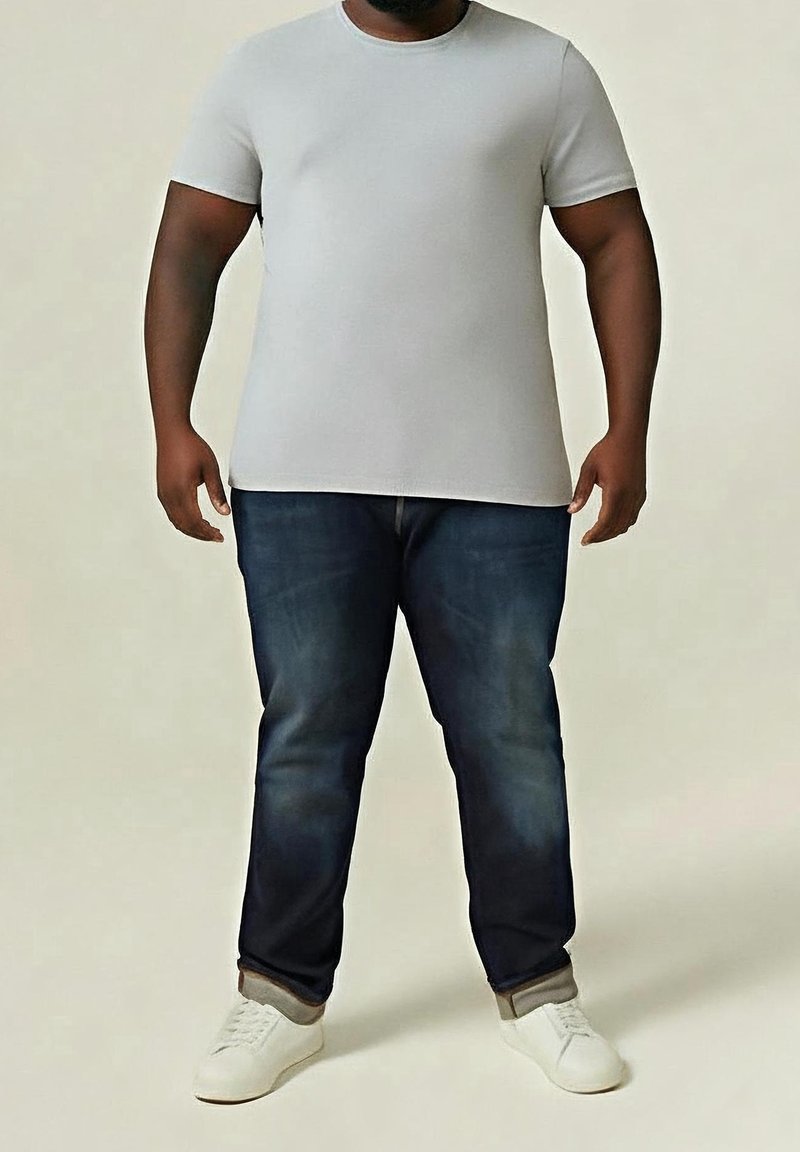 Homme portant un t-shirt blanc à manches courtes, un jean bleu foncé, des chaussettes beiges et des baskets blanches, debout sur un fond clair.