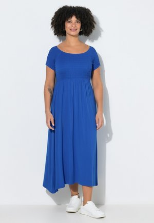 Frau mit lockigem Haar, die ein blaues Midikleid und weiße Turnschuhe trägt, steht vor einer weißen Wand mit einem schwachen Schatten.