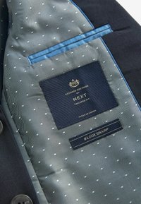 L'intérieur de la veste navy présente une doublure à pois blancs sur fond bleu clair. Comprend les étiquettes : "NEXT," "coupe ajustée," et "#LOOKSHARP."
