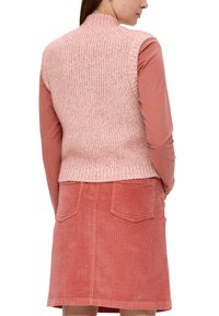 Frau in einem rosa gestrickten ärmellosen Pullunder über einem Langarmshirt und einem rosa Cordrock, von hinten gezeigt.