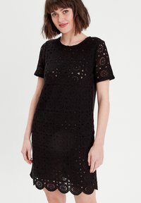 Robe en dentelle noire à manches courtes, avec des motifs découpés circulaires et un ourlet festonné. Tissu léger avec une finition texturée.