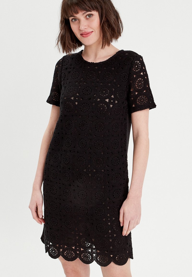 Robe en dentelle noire à manches courtes, avec des motifs découpés circulaires et un ourlet festonné. Tissu léger avec une finition texturée.